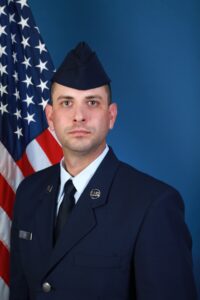 Dustin Aimes poses for his official U.S. Air Force service photo taken in August 2019, wearing his blue service coat, a light blue shirt and a blue flight cap, with the American flag behind his right shoulder.