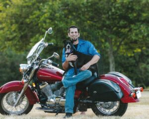 Dustin Aimes has a big smile on his face while sitting on his red Honda Shadow motorcycle and holding his dog, Lola, at the American River with trees in the background.