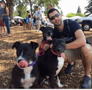 At a summertime outside event with popup tents and people in the background, Dustin Aimes enjoys time with his dogs Lexi, Lola and Sadie, who are all panting with their tongues out.