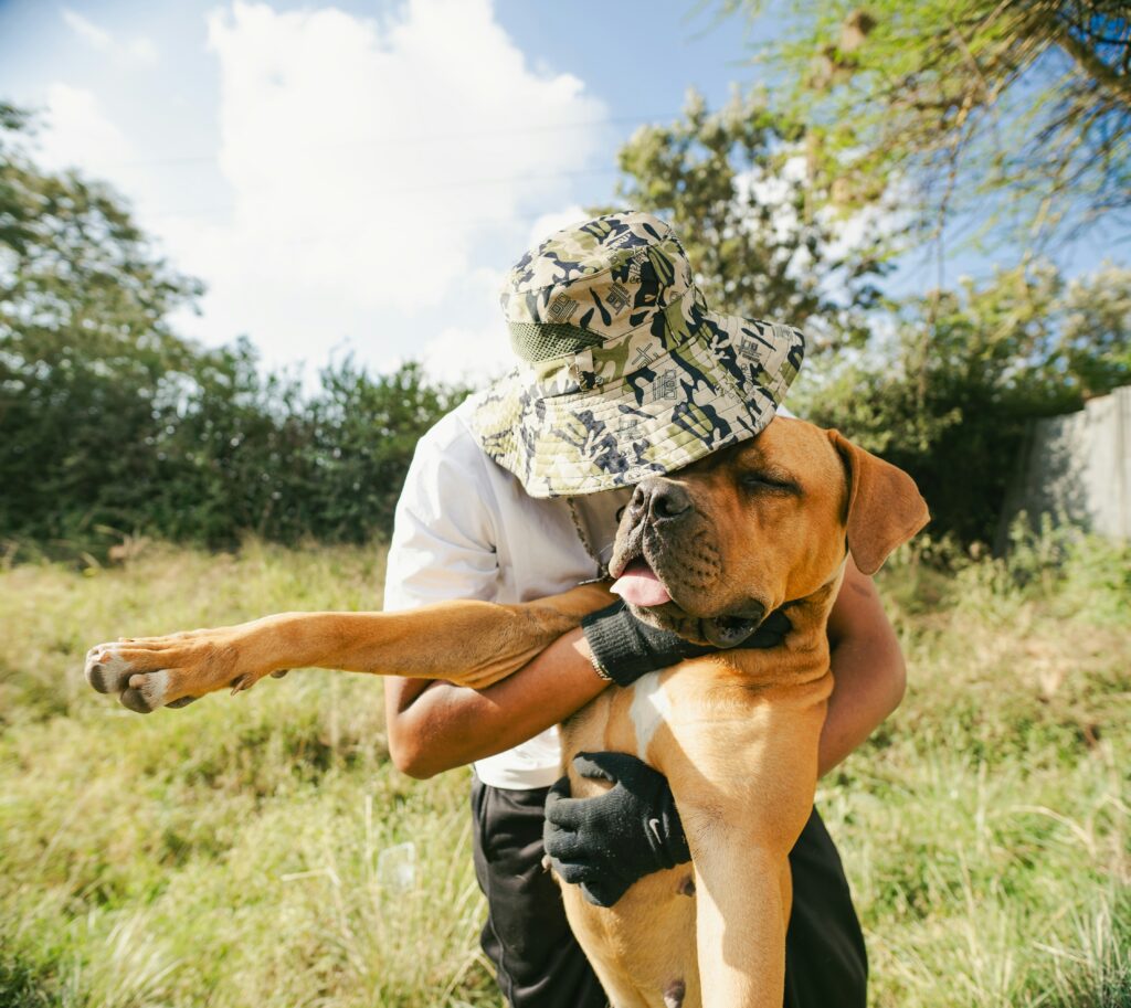 Wearing a camo hat, a man has his head down and is hugging a large tan dog that he is lifting up.